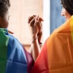 Black African LGBT woman covering LGBTQ rainbow flag with asian LGBT woman upside down, gay pride movement concept
