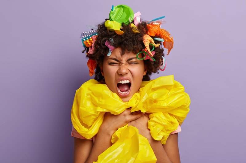 Headshot of irritated bothered young female wrapped in polythene sheeting, screams loudly, smirks face, keeps mouth widely, isolated on purple background. Stop using plastic. Recycling concept