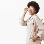 Backpack always ready for adventures. Stylish good-looking female tourist with dark skin and afro hairstyle checking glasses on eyes and turning at camera while standing in profile over gray wall