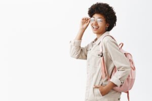 Backpack always ready for adventures. Stylish good-looking female tourist with dark skin and afro hairstyle checking glasses on eyes and turning at camera while standing in profile over gray wall