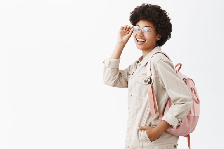 Backpack always ready for adventures. Stylish good-looking female tourist with dark skin and afro hairstyle checking glasses on eyes and turning at camera while standing in profile over gray wall